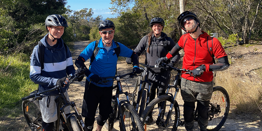 A group of 4 people in bike riding attire, standing together with their bikes in the bush